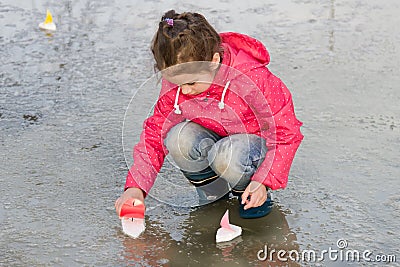 Happy cute little girl in rain boots playing with handmade colorful ships in the spring water puddle Stock Photo