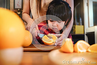 Happy Cute 3-4 Years Old Girl with her Mom Slice some Orange on Wooden Table in Pantry Room. Young Girl is Learning Cook with her Stock Photo