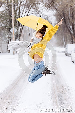 Happy dark-haired girl dressed in a yellow sweater, jeans and a white scarf is jumping with a yellow umbrella in a snowy Stock Photo