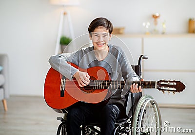 Happy disabled teen boy in wheelchair playing guitar, using musical instrument indoors. Teenage hobbies and pastimes Stock Photo