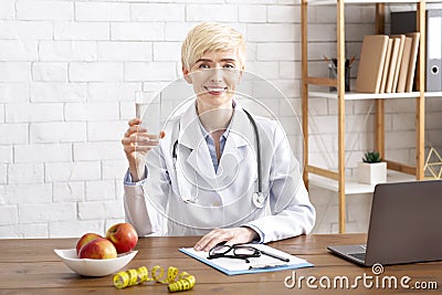 Happy doctor nutritionist looks at camera, holds glass of water, sits at table with apples on plate Stock Photo