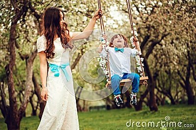Happy dressy mother and toddler child son having fun on swing in spring or summer park Stock Photo