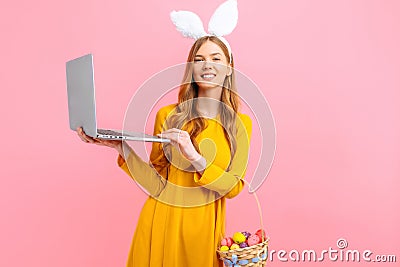 Happy Easter. Happy woman in the ears of an Easter Bunny holding a basket of Easter eggs using a laptop on an pink background Stock Photo