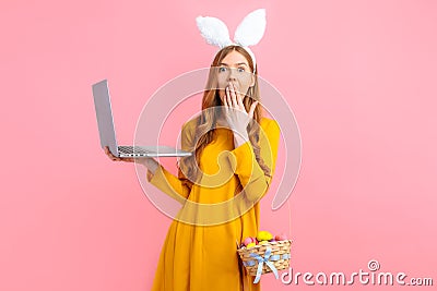 Happy Easter. A shocked woman in the ears of an Easter Bunny holding a basket Easter eggs using a laptop on an pink background Stock Photo