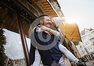 Happy enamored couple is standing outdoors near the decorated shop windows, hugging and kissing each other Stock Photo