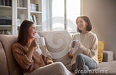 Happy european young women in casual with cups drink coffee, communication, sit on sofa in living room Stock Photo