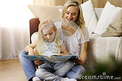 Happy family. Blonde young mother reading a book to her cute daughter while sitting at wooden floor in sunny room Stock Photo