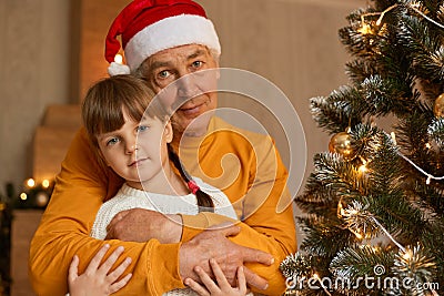 Happy family on Christmas Eve, little charming little girl with pigtails posing with her grandfather, senior man in santa claus Stock Photo