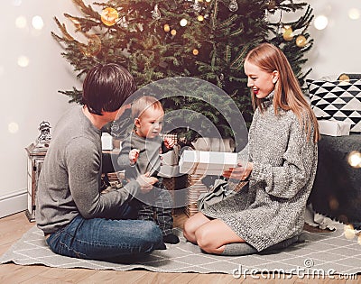 Happy family couple give gifts in the living room, behind the decorated xmas tree, the light give a cozy atmosphere. New Year Stock Photo