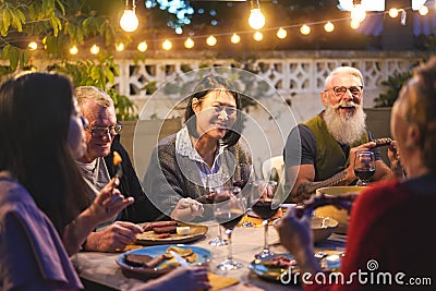 Happy family eating and drinking wine at barbecue dinner outdoor - Multiracial mature and young people having fun at bbq sunday Stock Photo