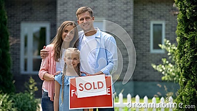 Happy family holding sold sign, standing against new bought house, thumbs up Stock Photo