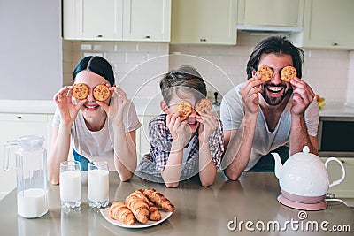 Happy family are playing with rolls. They pyt them on eyes and smiling. Family are leaning to table. There are kettle Stock Photo