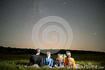 Happy family resting in night field looking at dark sky with many bright stars. Parents and children observing meteor shower Stock Photo