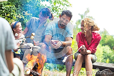 Happy family sitting with woman while roasting marshmallows over burning campfire at park Stock Photo