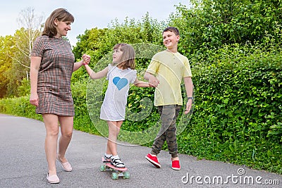 A happy family teaches a child to ride a skateboard in a park. Mom with children plays outdoors Stock Photo