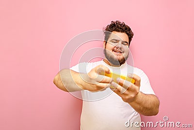 Happy fat gamer playing mobile games on smartphone with smile on face on pink background. Curly overweight kid in white t-shirt Stock Photo