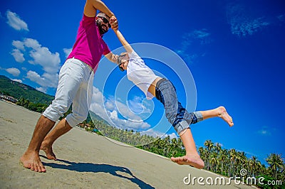 Happy father is spinning in circle his adorable boy. Sunny tropical beach, palm trees behind Stock Photo