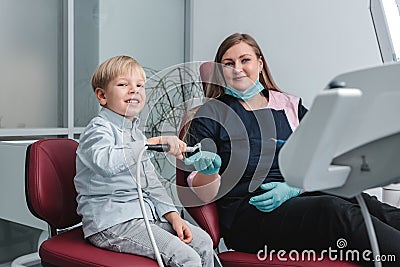 Happy female dentist sitting in dental chair and showing to patient how not to fear working drill, young boy holding the tool and Stock Photo