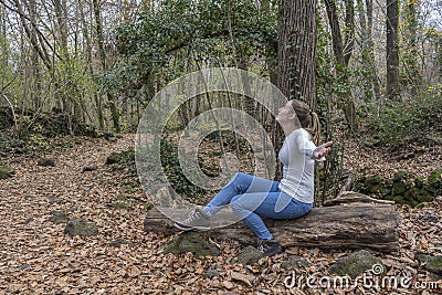 Happy female with open arms and stylish jeans fashion sitting on a tree in the forest.Celebration and happiness concept.Travel Stock Photo