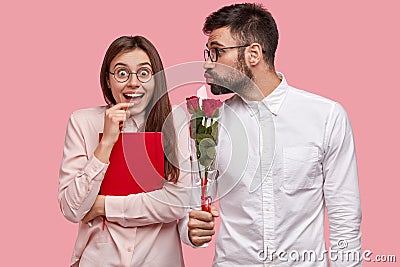 Happy female student in spectacles, carries red textbook, glad to recieve bouquet of roses from male wonk Stock Photo