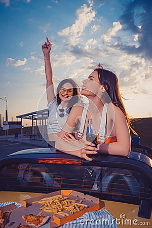 Happy females having fun and drinking soda in glass bottles, posing in yellow car with french fries and pizza on its Stock Photo