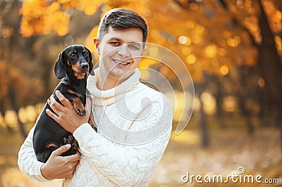 Happy free time with beloved dog! Handsome young man staying in autumn park smiling and holding cute puppy dachshund. Stock Photo