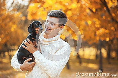 Happy free time with beloved dog! Handsome young man staying in autumn sunny park smiling and holding cute puppy dachshund Stock Photo