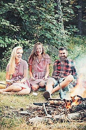 Happy friends at campfire. Bearded man and women smile at bonfire. Hipster in palid shirt and girls in vintage dresses Stock Photo