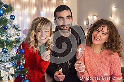 Happy friends holding sprinklers by the Christmas tree on new years eve, celebrating and smiling Stock Photo