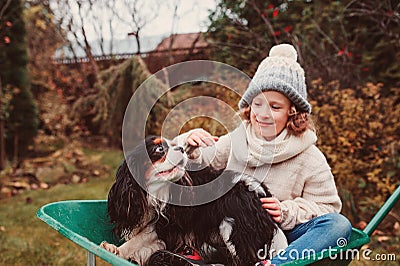 Happy funny child girl riding her dog in wheelbarrow in autumn garden, candid outdoor capture Stock Photo