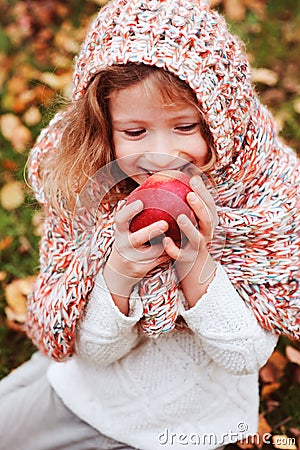 Happy funny kid girl in cozy knitted scarf eating fresh apple in autumn garden Stock Photo