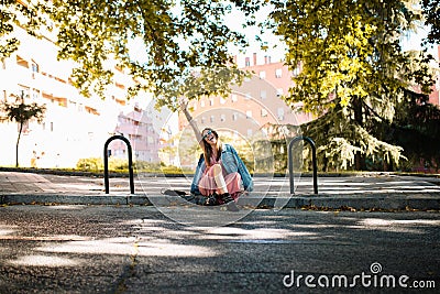 Happy and funny young woman with sunglasses sitting on the sidewalk raising her hand and waiting for someone in the park. Human Stock Photo