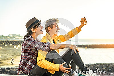 Happy gay couple taking a selfie with mobile smart phone camera on the beach at sunset - Lesbians having fun in holiday Stock Photo