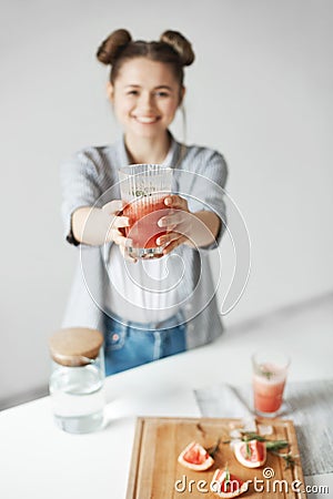 Happy girl with buns smiling stretching grapefruit detox smoothie to camera over white wall. Healthy diet food. Focus o Stock Photo