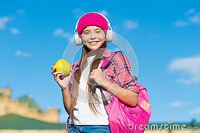 Happy girl in casual fashion style listen to music in modern headphones holding apple fruit for travel sunny outdoors Stock Photo