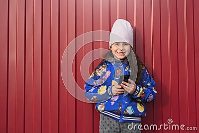 Happy girl kid with smartphone in hands on a red background in the street. Dressed in a blue jacket and smiling. Stock Photo