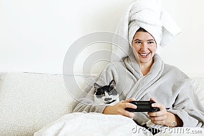Happy girl playing video games with cat in bed. Beautiful young woman in bathrobe relaxing and lying in bed , hugging cat and Stock Photo
