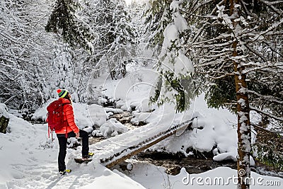 A happy girl, with a red jacket and a red backpack, crossing a wooden bridge, during a winter hiking trip Stock Photo