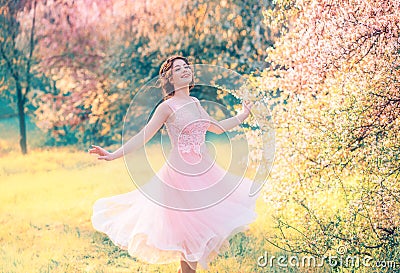 Happy girl in short flying gentle pink dress laughs joyfully, doll princess whirls in bright yellow spring garden with Stock Photo