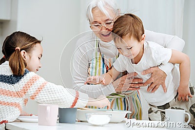 Happy granny holding her grandson, standing on a stool, reaching for the food Stock Photo