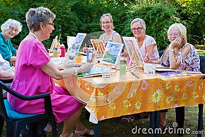 Happy group of senior ladies enjoying art class seated around a table outdoors in the garden painting with water colors while Stock Photo