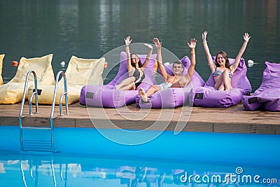 Happy guy and two attractive girls sitting on a cushioned lounger with his hands raised up and looking at the camera Stock Photo