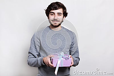 Happy handsome bearded guy in grey sweater holding gift and smiling to camera on white background. Holidays time, making surpris Stock Photo