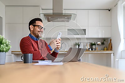 Happy handsome businessman messaging online on smart phone while using laptop on desk at home Stock Photo