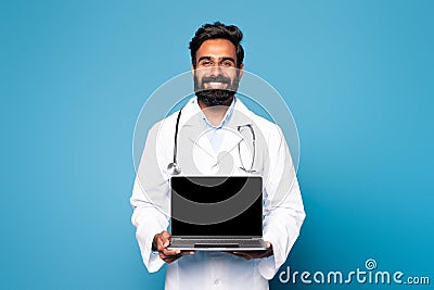Happy hindu man doctor in white coat with stethoscope showing laptop with blank screen on blue studio background Stock Photo