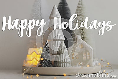 Happy Holidays text on stylish little Christmas trees and house on white table. Season`s greeting card. Cozy christmas magical Stock Photo