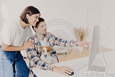 Happy husband points into monitor of computer, asks for useful advice in wife what better to buy, make shopping online, pose at Stock Photo