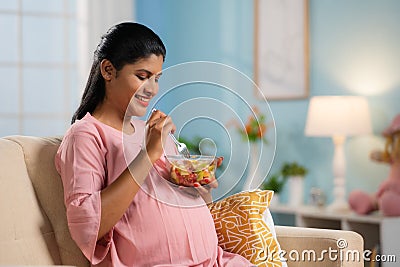 Happy indian pregnant woman eating fruit salad while sitting on sofa at home - concept of Pregnancy nutrition, Healthy Stock Photo