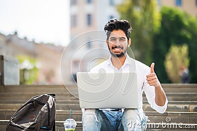 Happy indian student sitting on the stairs showing thumb up working on laptop, in the university campus. Technology, education and Stock Photo