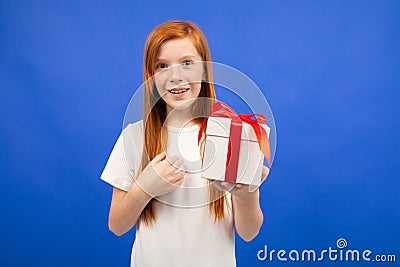 Happy joyful teenager girl with red hair does not believe happiness holding a gift box on a blue studio background Stock Photo